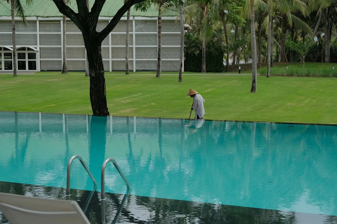 Operario de mantenimiento limpiando con una pértiga el borde de una piscina desbordante de agua turquesa, junto a una zona ajardinada