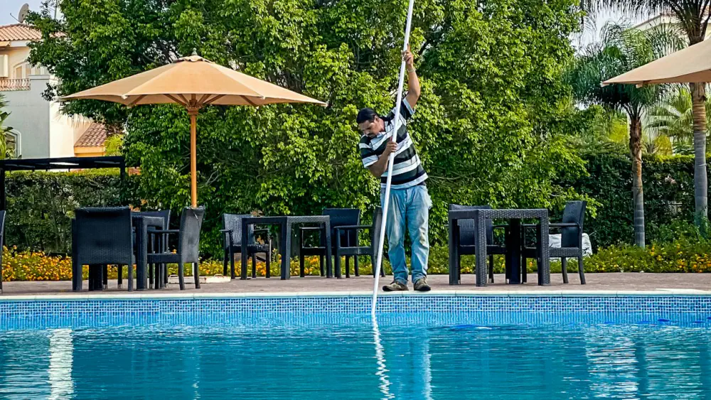 Operario de mantenimiento limpiando con una pértiga telescópica el fondo de una piscina de agua limpia y transparente en una zona ajardinada