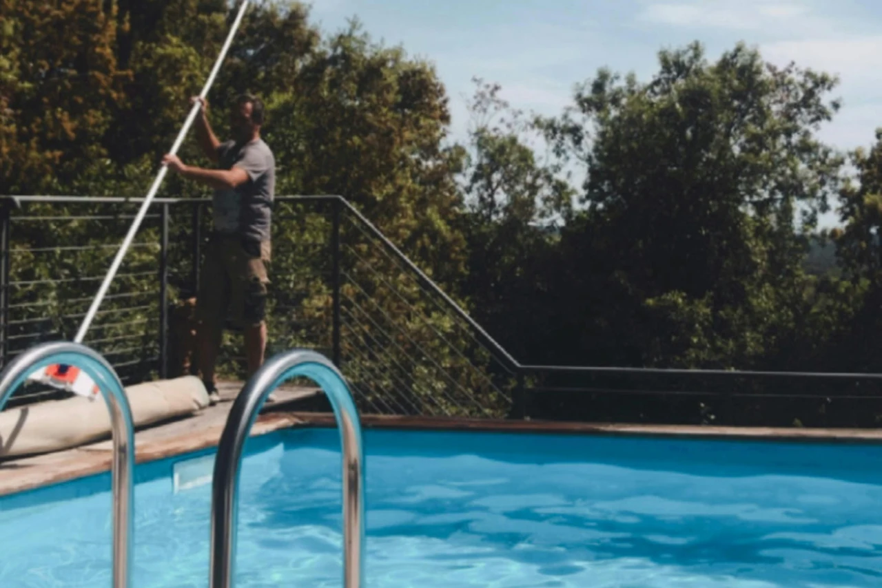 Operario de mantenimiento manejando una pértiga telescópica junto a una piscina residencial de agua limpia, con escalera de acero inoxidable y tarima de madera